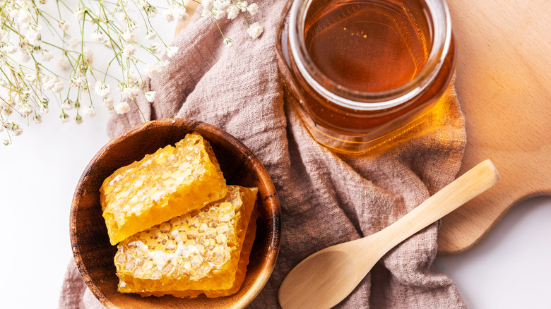 A close-up of an open glass jar of honey on a coarse cloth and a wooden cutting board, with flowers, a small wooden spoon, and some honeycombs in a wooden bowl to the side