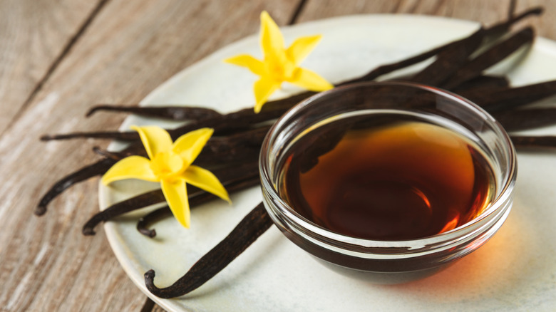 Dish of vanilla extract next to vanilla bean pods and flowers
