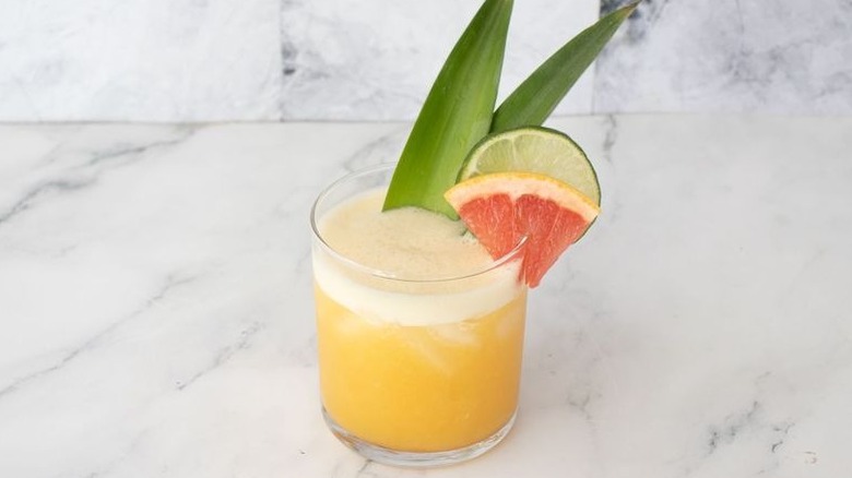 Yellow cocktail garnished with pineapple leaves, lime slice, and grapefruit slice