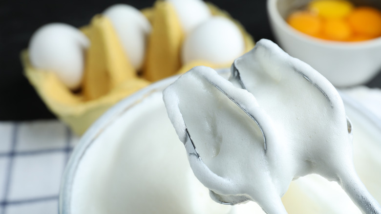 A close up of foamy egg whites on a hand-mixer with fresh eggs and a bowl of egg yolks in the background