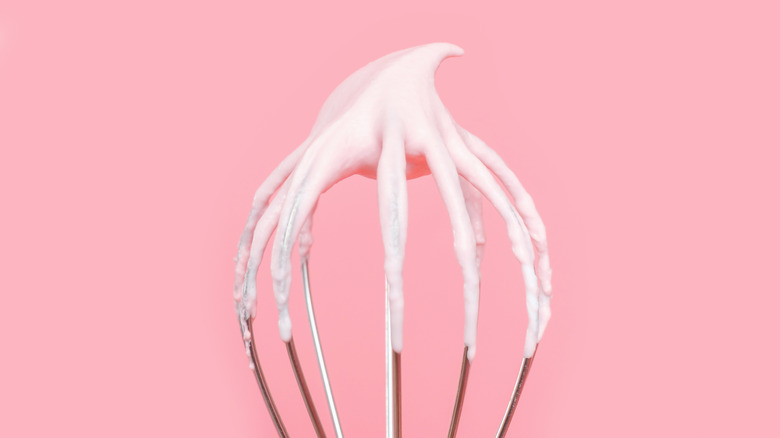 An upside down whisk showing egg whites whipped to stuff peaks, against a pink background