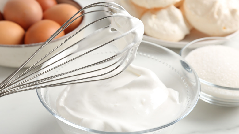 A glass bowl of homemade whipped cream with a whisk above it, implying that it was used to whip said cream