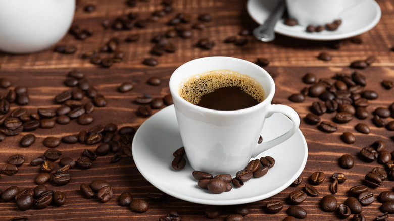 A small white mug of coffee sitting on a small white plate surrounded by coffee beans