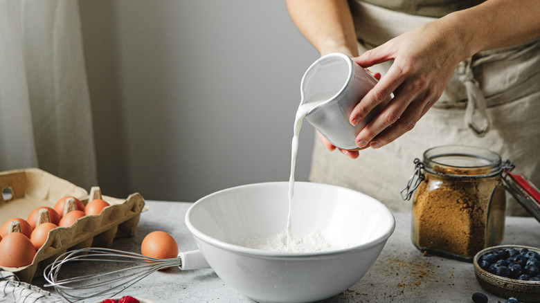 Someone pouring milk into a bowl with flour to make baked goods, with eggs and other ingredients nearby