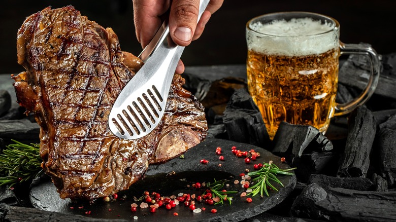 Person holding steak in tongs next to glass of beer