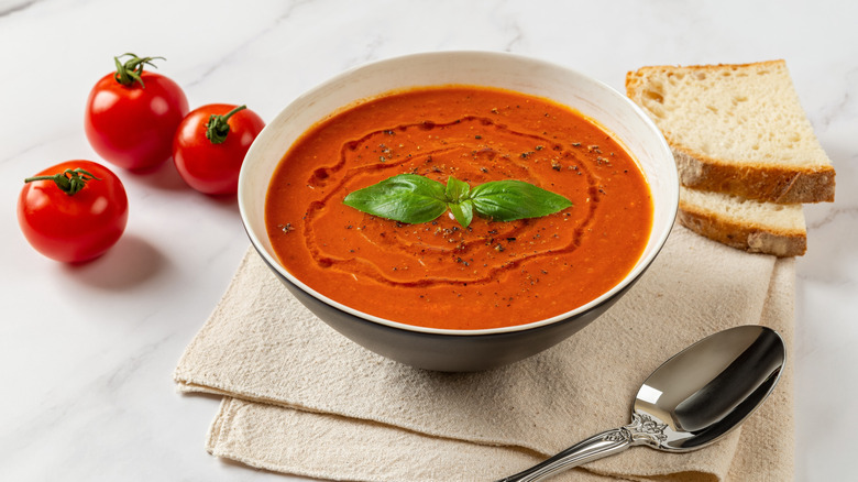 A bowl of hot tomato soup next to tomatoes and sliced bread.